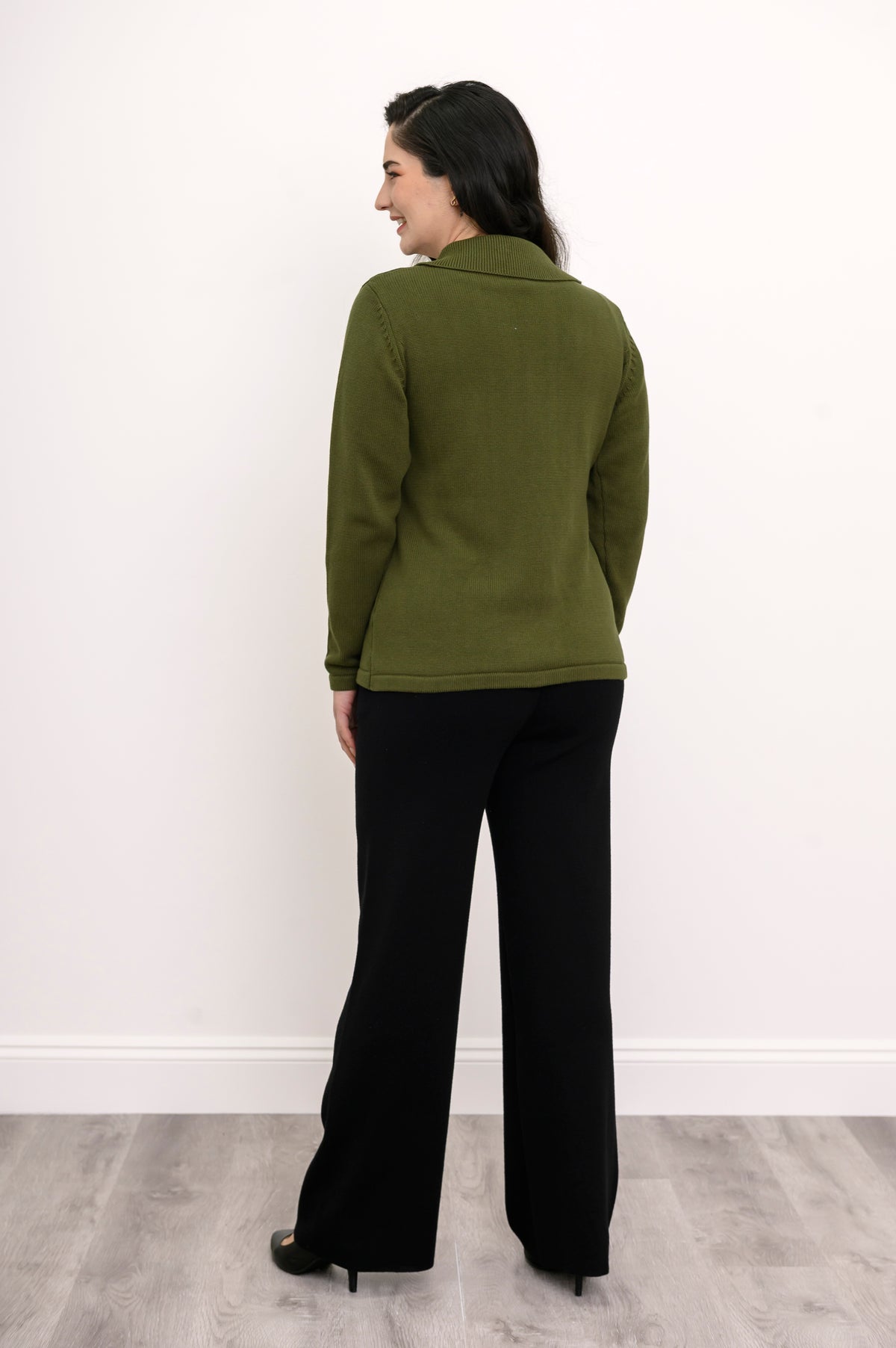 Back view of a woman wearing a hip length green cotton sweater highlighting the clean tailoring and shoulder fit.