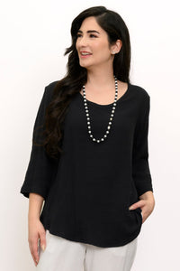 Woman wearing a black top and white pants with a beaded necklace against a white wall, front view