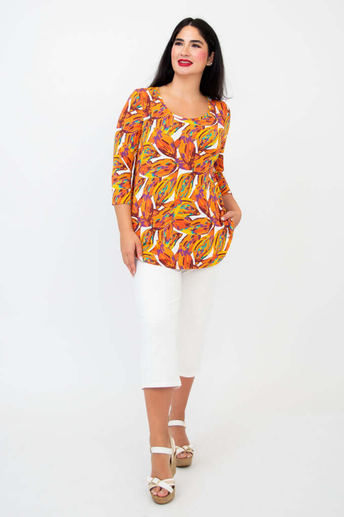 Woman in a bold orange leaf print top paired with white capri pants, standing and smiling