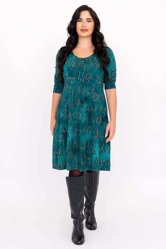 Front view of model wearing teal A-line dress with petroglyph pattern and black boots