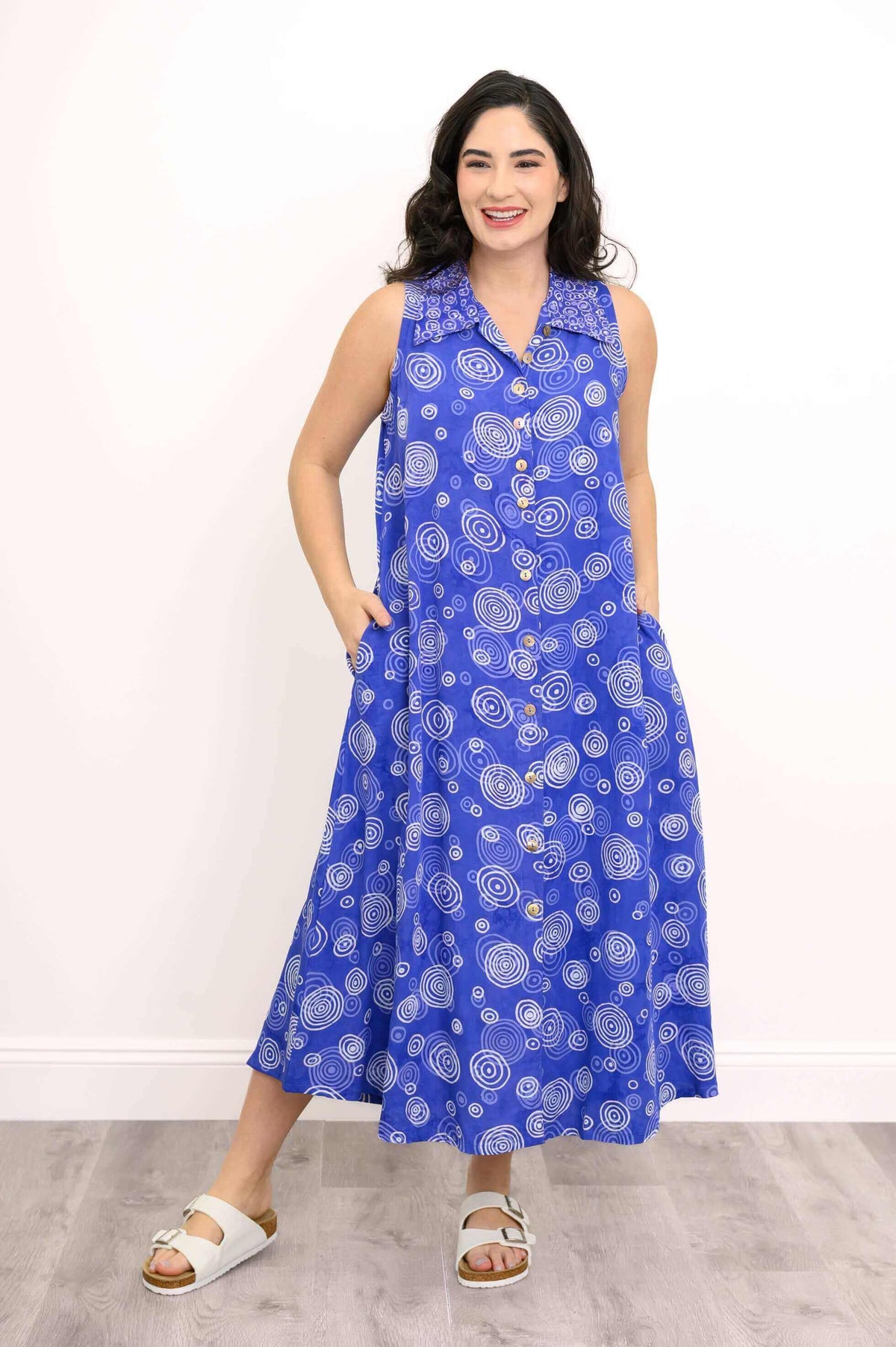 Model wearing blue A-line sleeveless dress, facing forward with hands in pockets