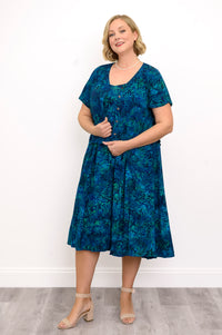 Woman wearing a blue patterned dress and blue patterned blouse standing against a white wall, front view