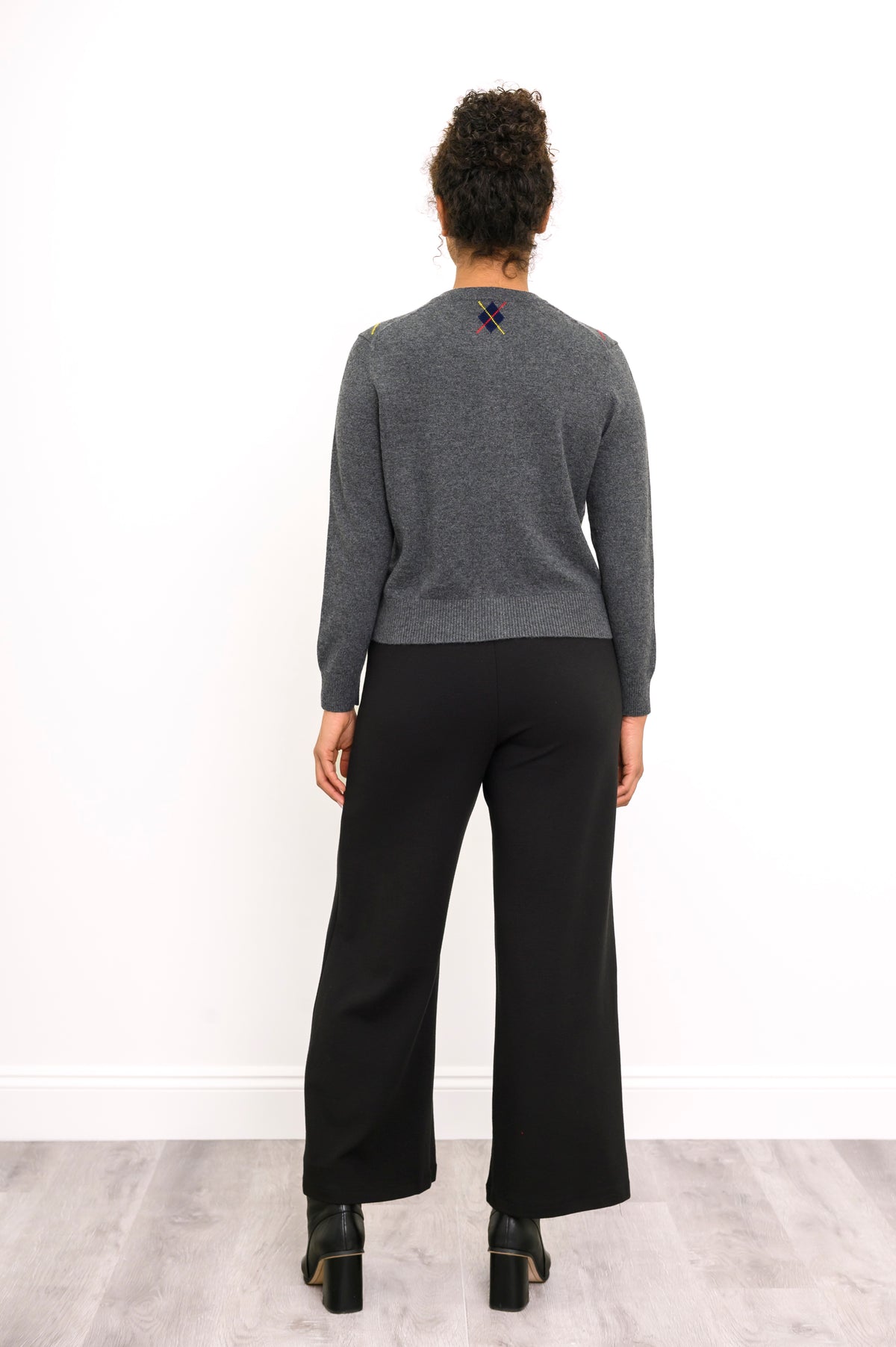Back view of woman standing wearing a grey knit sweater with a small argyle emblem near the neck