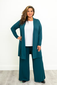 Full-length portrait of a smiling woman wearing an open crystal teal long cardigan over a white shirt and teal pants.
