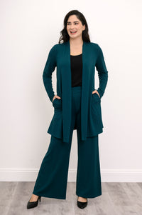 Wide studio shot of a loose-fit long teal cardigan styled with a monochrome knit top and trousers.