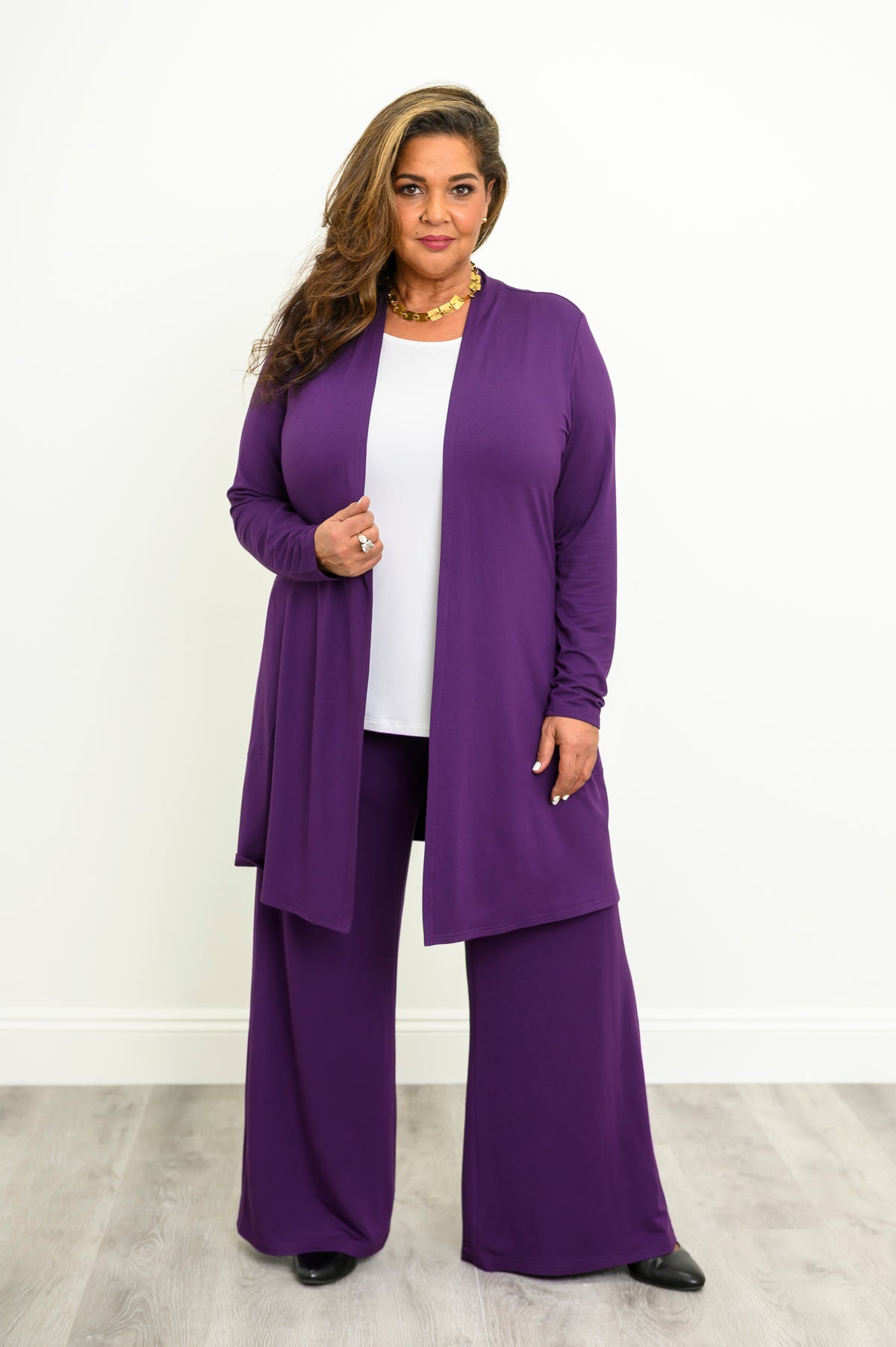 Woman wearing long purple cardigan over a white top and purple pants on a white background, front view