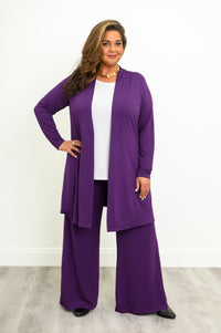 Woman wearing long purple cardigan over a white top and purple pants on a white background, front view