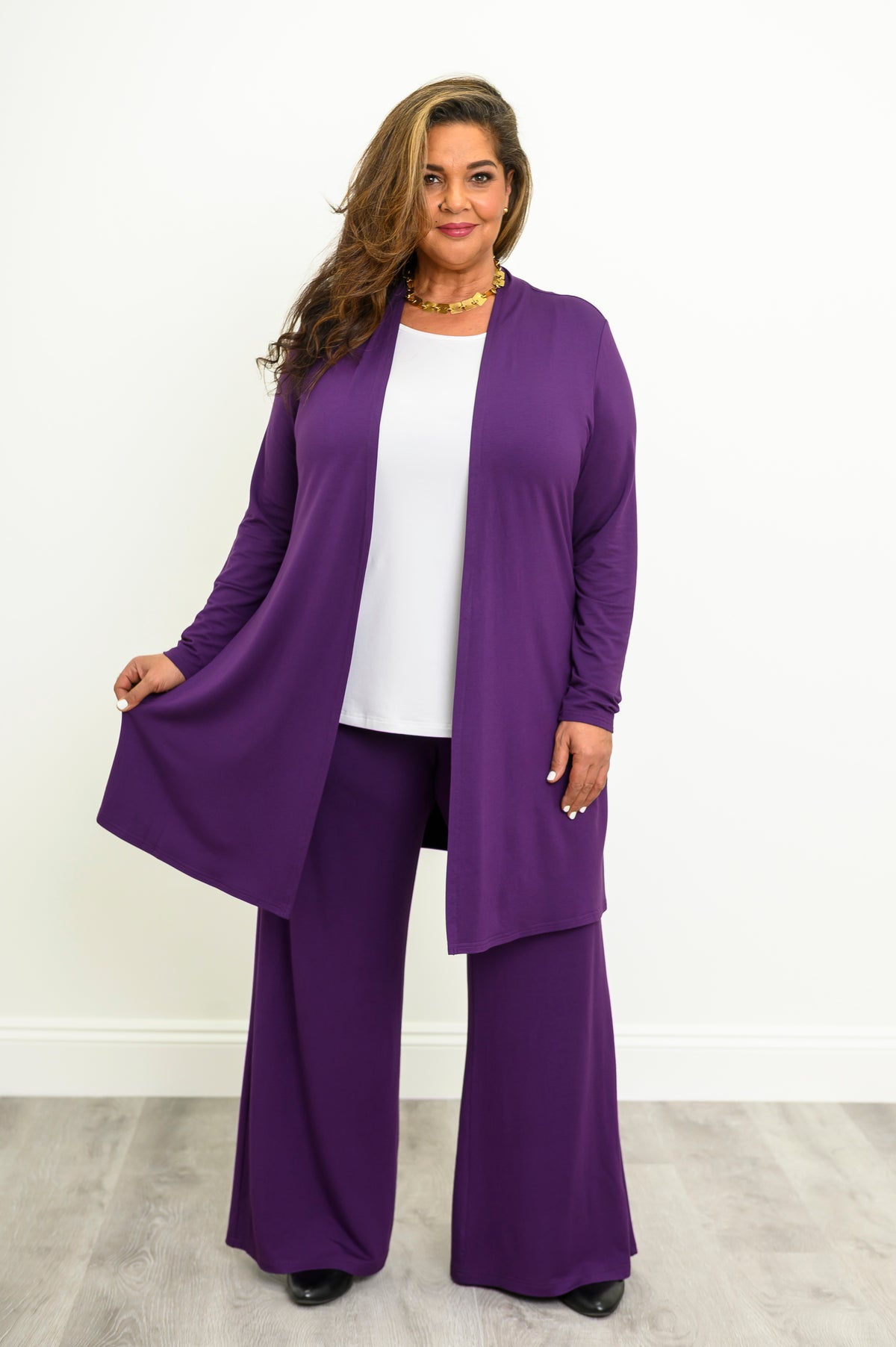 Woman wearing long purple cardigan over a white top and purple pants on a white background, front view