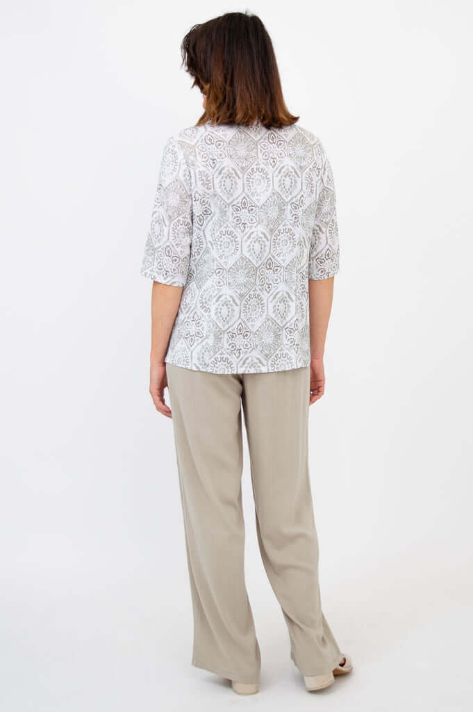Back view of a woman in a patterned blouse with beige pants and sandals, standing against a white background