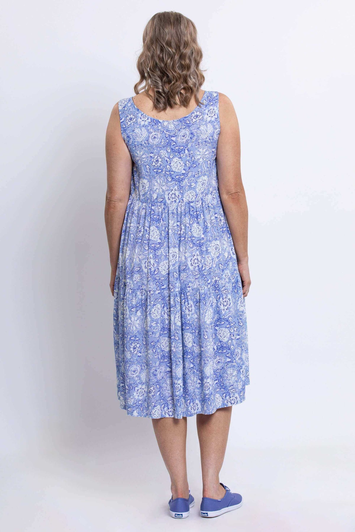 Back view of a woman with wavy hair wearing a sleeveless blue floral dress and blue sneakers