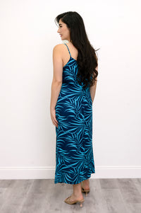 Back view of woman wearing a blue leaf-patterned dress and beige sandals against a white background