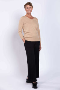 Woman in beige V-neck sweater and black pants with hands in pockets, smiling