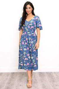 A woman modeling a short-sleeve midi dress with a blue, pink, and white tropical floral print, standing against a white wall and light gray wood floor.