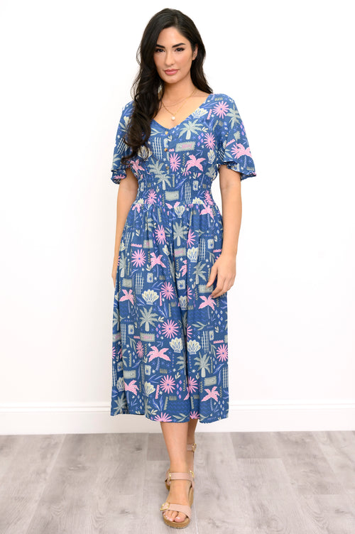 A woman modeling a short-sleeve midi dress with a blue, pink, and white tropical floral print, standing against a white wall and light gray wood floor.