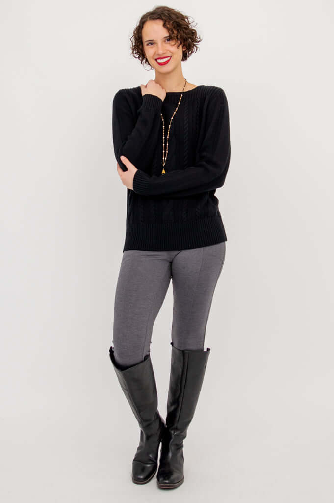 Woman standing in black cable knit sweater with arms folded, wearing grey pants and tall boots