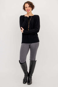 Woman standing in black cable knit sweater with arms folded, wearing grey pants and tall boots
