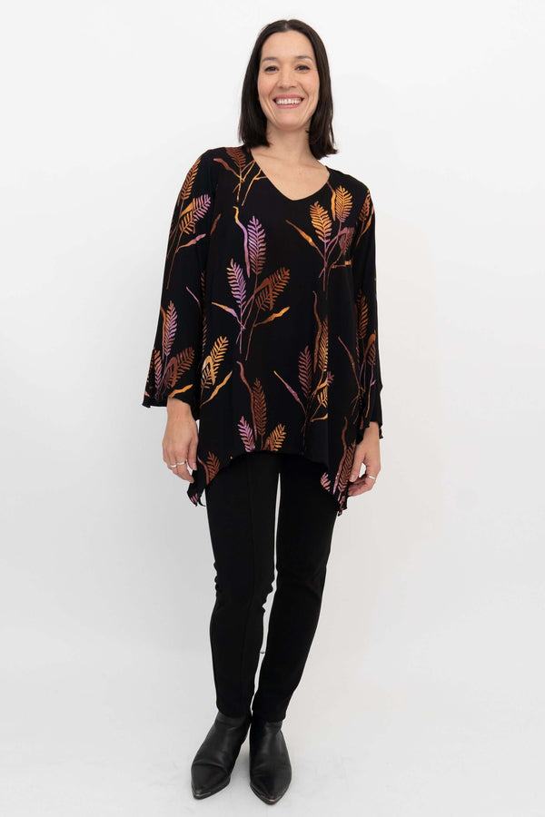 Full body view of a woman in a black shark-bite hem top with autumn leaf pattern and black pants