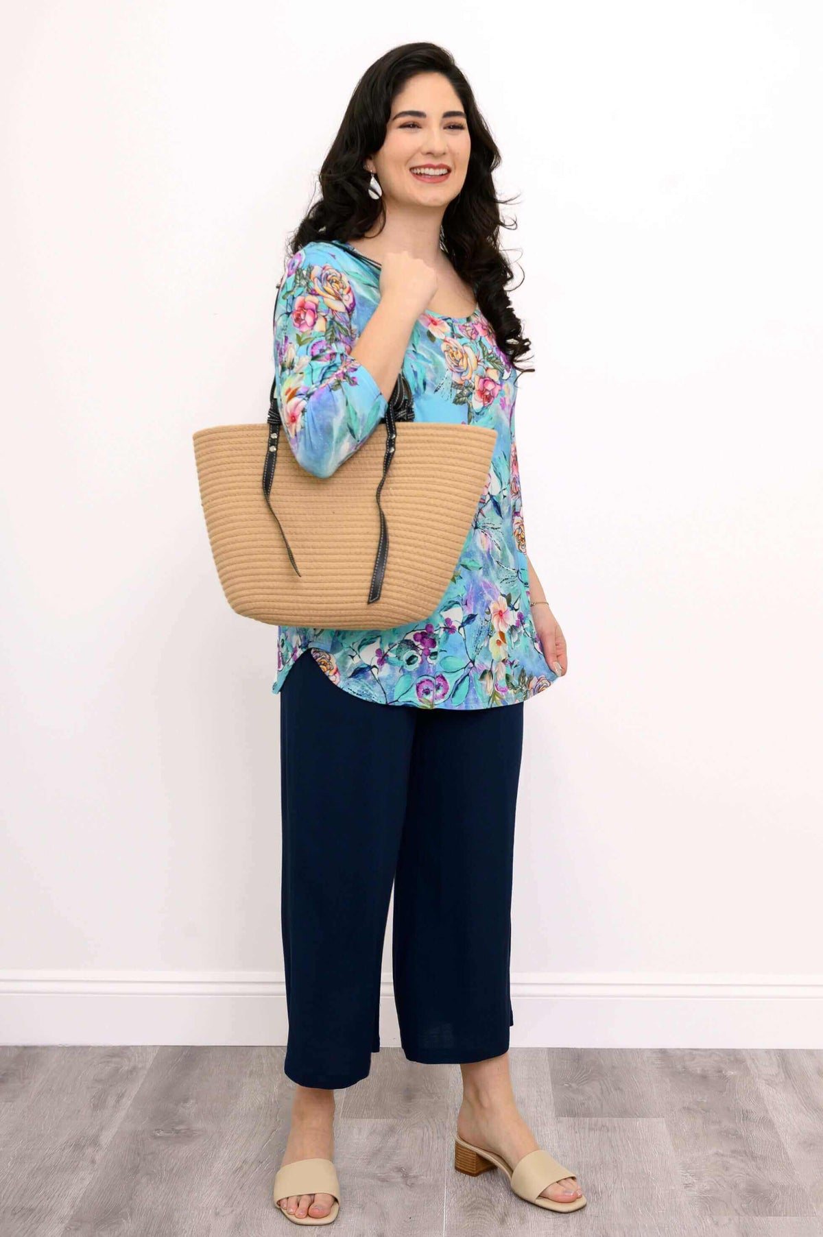 Woman in blue floral top with navy pants and tan purse, standing