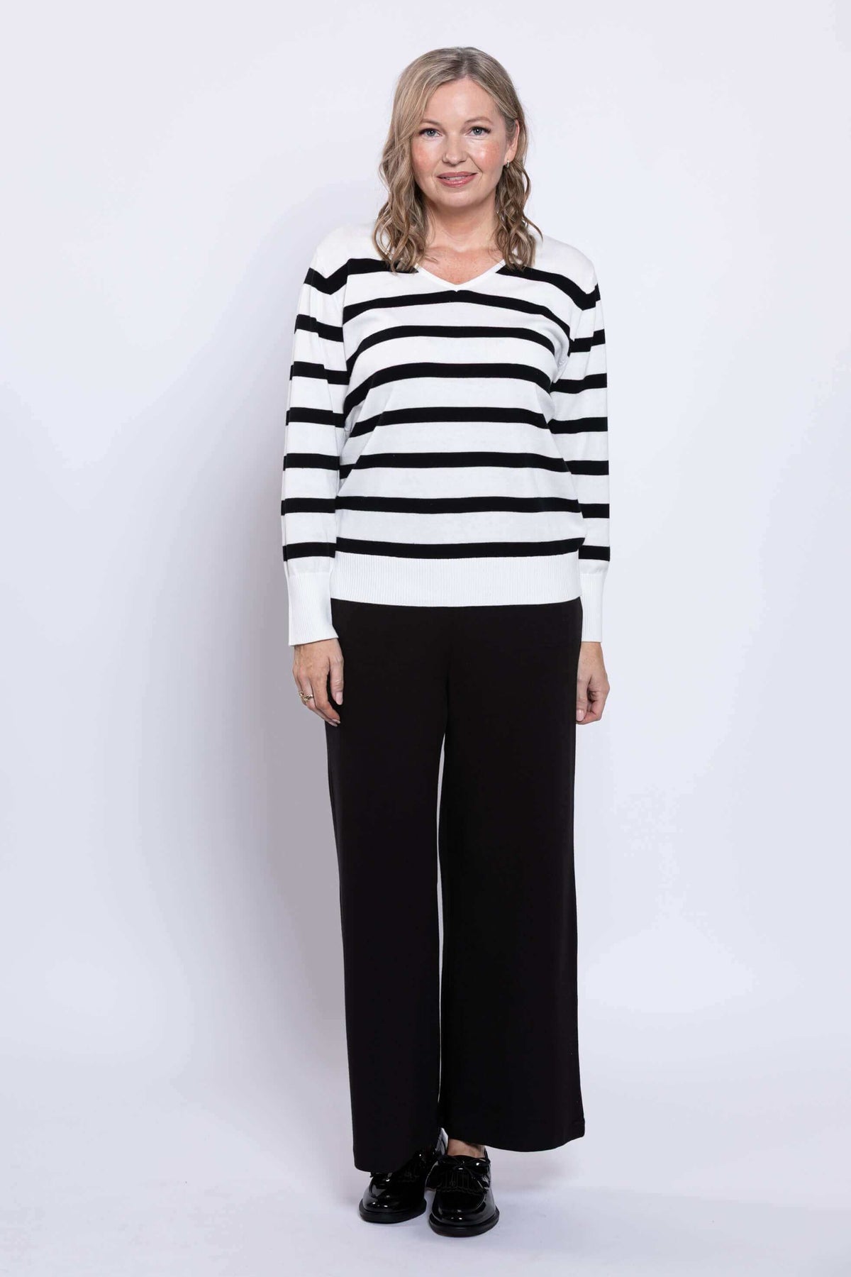 Full body view of a woman in a black and white Breton stripe V-neck sweater and black pants