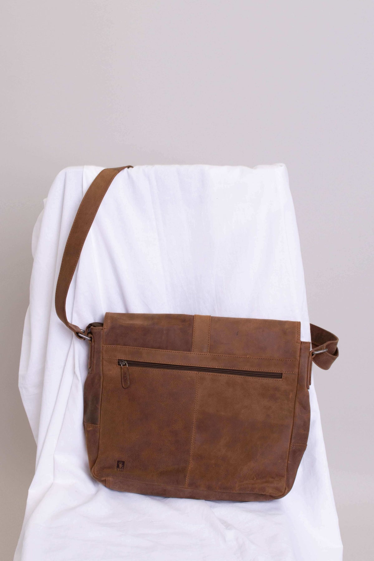 Brown buffalo leather messenger bag displayed on a draped white chair, back zipper visible