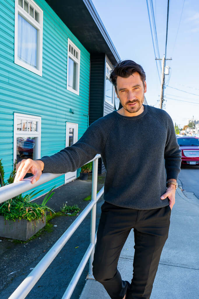 Man in charcoal sweater leaning casually on a rail outdoors by a teal building