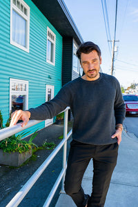 Man in charcoal sweater leaning casually on a rail outdoors by a teal building