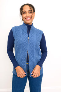 Model in a relaxed, open blue cable knit vest layered over a dark top and matching denim jeans, standing front view.