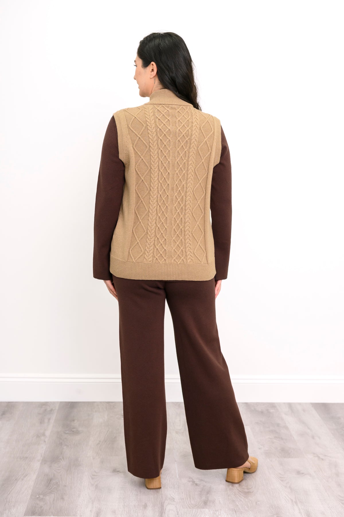 Back view of a honey-beige cable knit vest, highlighting the vertical sweater texture and straight, ribbed hemline.