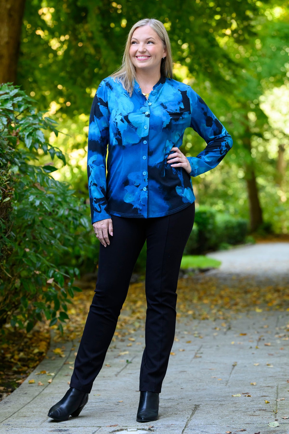 Front view of a female model wearing a blue patterned buttoned-up long-sleeve blouse with black pants and black boots, against a background of sunlit trees and a path