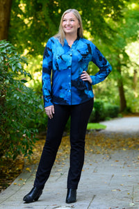 Front view of a female model wearing a blue patterned buttoned-up long-sleeve blouse with black pants and black boots, against a background of sunlit trees and a path
