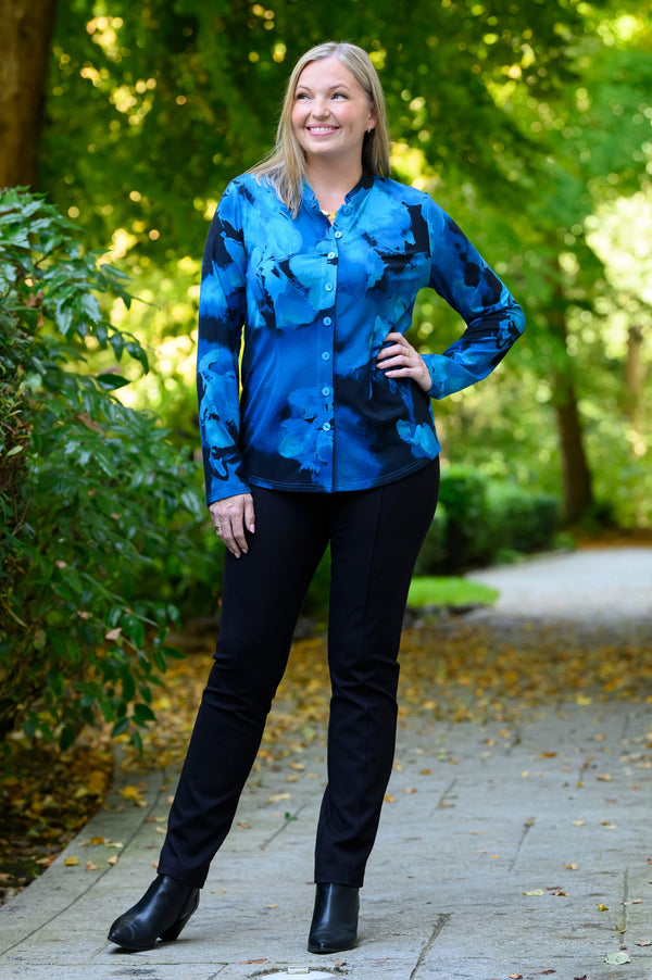 Front view of a female model wearing a blue patterned buttoned-up long-sleeve blouse with black pants and black boots, against a background of sunlit trees and a path