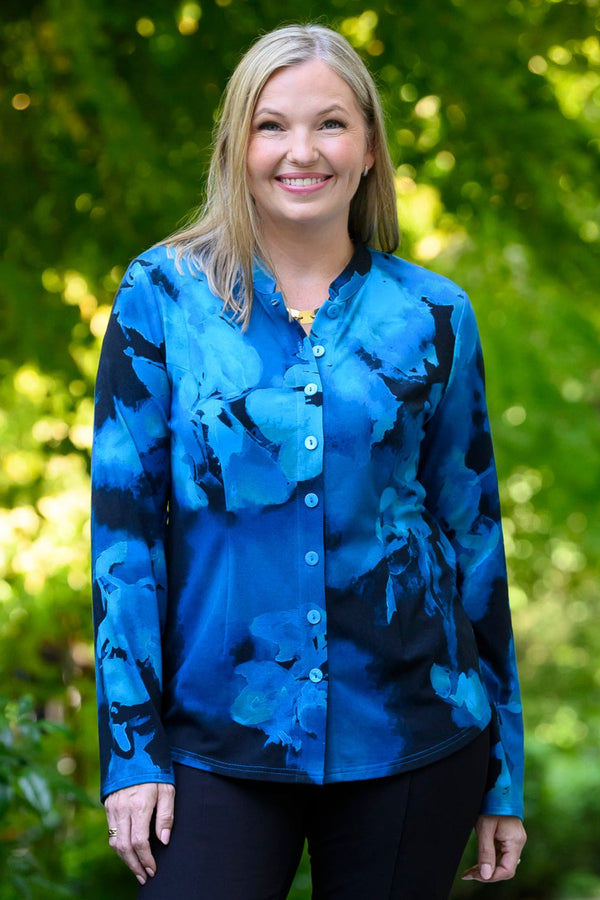 Front view of a female model wearing a blue patterned buttoned-up long-sleeve blouse with black pants, against a background of sunlit trees