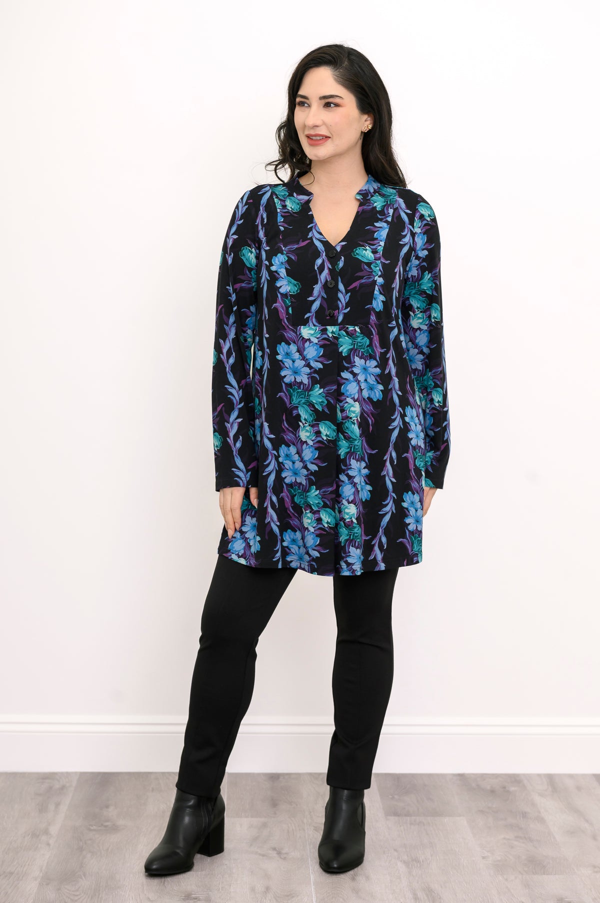 Model in a patterned teal button-up collar blouse and black pants, posing against a plain white wall.