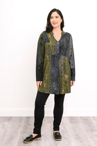Model in an olive-grey geometric print long-sleeve tunic with black trousers, standing against a white wall.