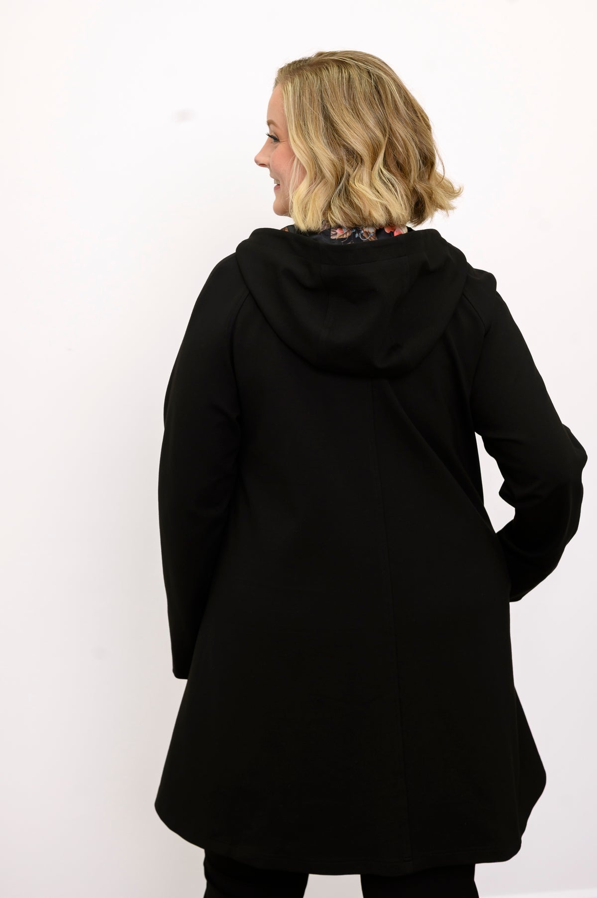 Rear profile shot of a long sleeve black swing coat displaying fluid drape and hooded back seam.