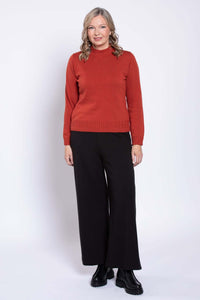 Woman standing in a rust colored mock neck sweater and black trousers, facing forward