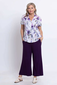 Front three-quarter view of a woman in a floral button-up shirt and purple pants