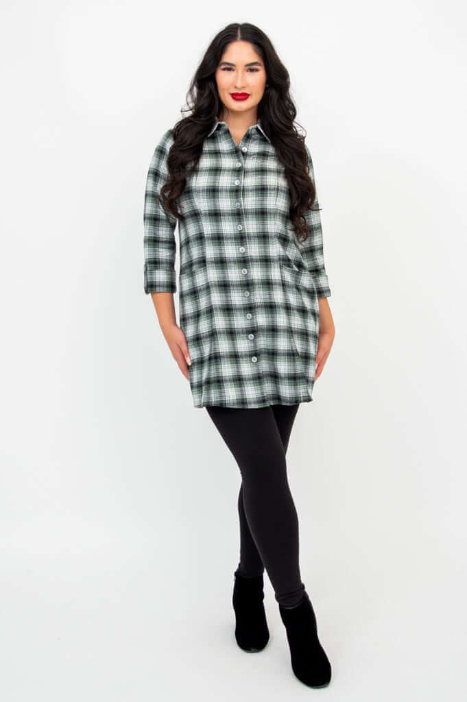 Full-body view of a woman in a green plaid tunic shirt with black leggings and boots
