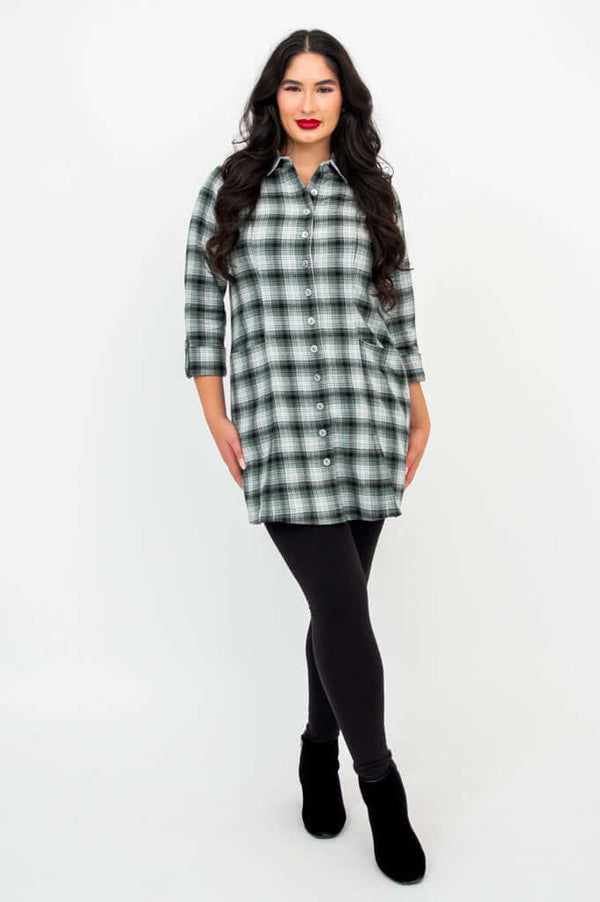 Full-body view of a woman in a green plaid tunic shirt with black leggings and boots