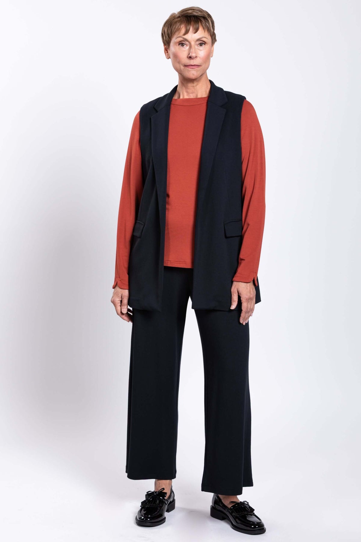Full body view of a woman in a black open vest, rust top, and black pants