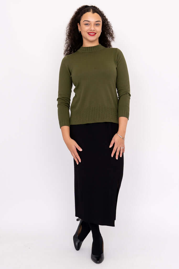 Full view of woman in olive green sweater and black skirt, standing against white background