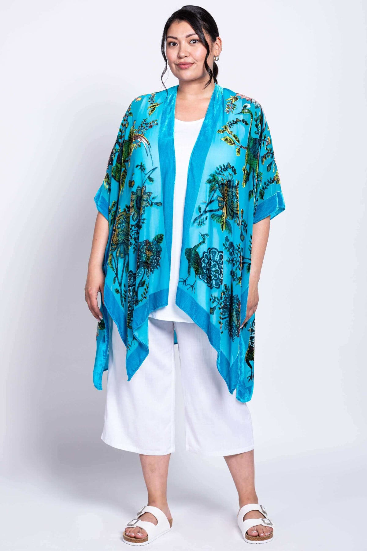 Model in full view wearing turquoise kimono wrap with floral prints, styled with white pants
