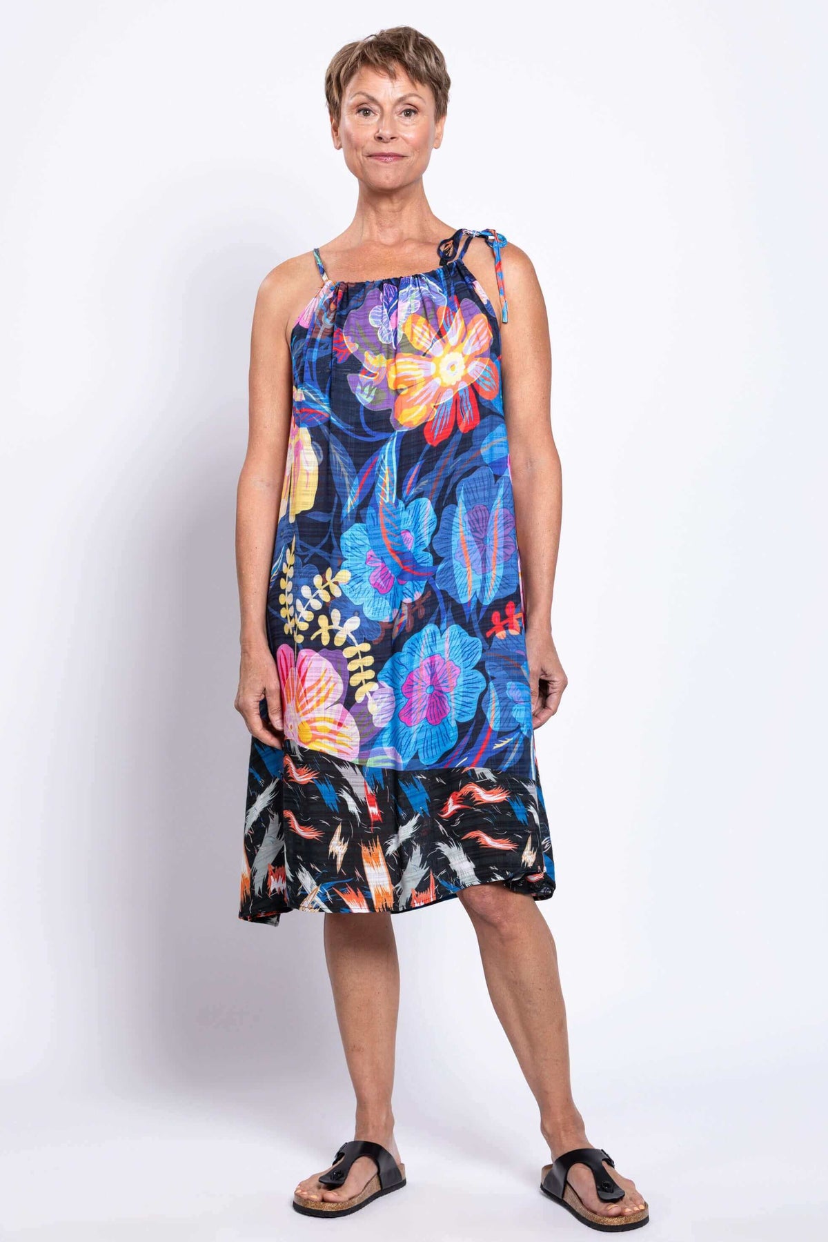 Full body front view of a woman in a vibrant sleeveless floral summer dress with one tie strap