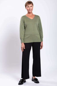 Woman standing in a sage green V-neck sweater and black pants, hands at sides