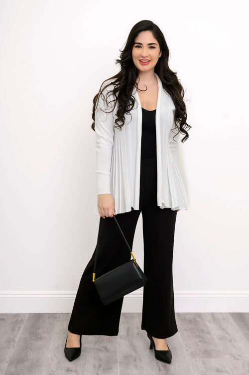 Woman standing in a white cardigan with pleated back, black top and pants, holding black purse