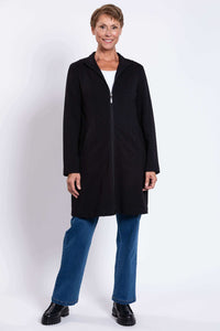 Full view of woman in black zip-front coat and blue jeans, standing and smiling