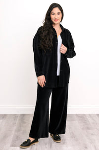 A woman modeling a black velvet two-piece lounge set consisting of a button-up shirt jacket and matching wide-leg pants, styled over a white top and black loafers.