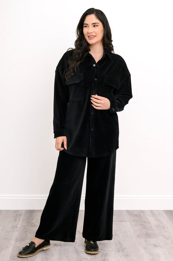 A woman modeling a black velvet button-up shirt and matching wide-leg pants set, paired with black slip-on shoes, against a white wall and light gray wood floor.
