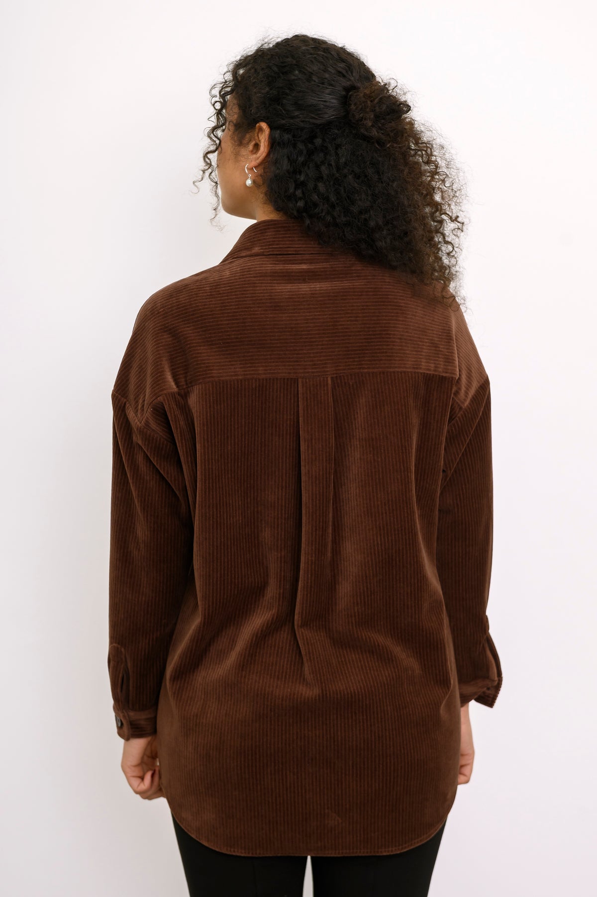 Back view of an oversized dark brown corduroy shirt jacket featuring a pleated back yoke and curved hem