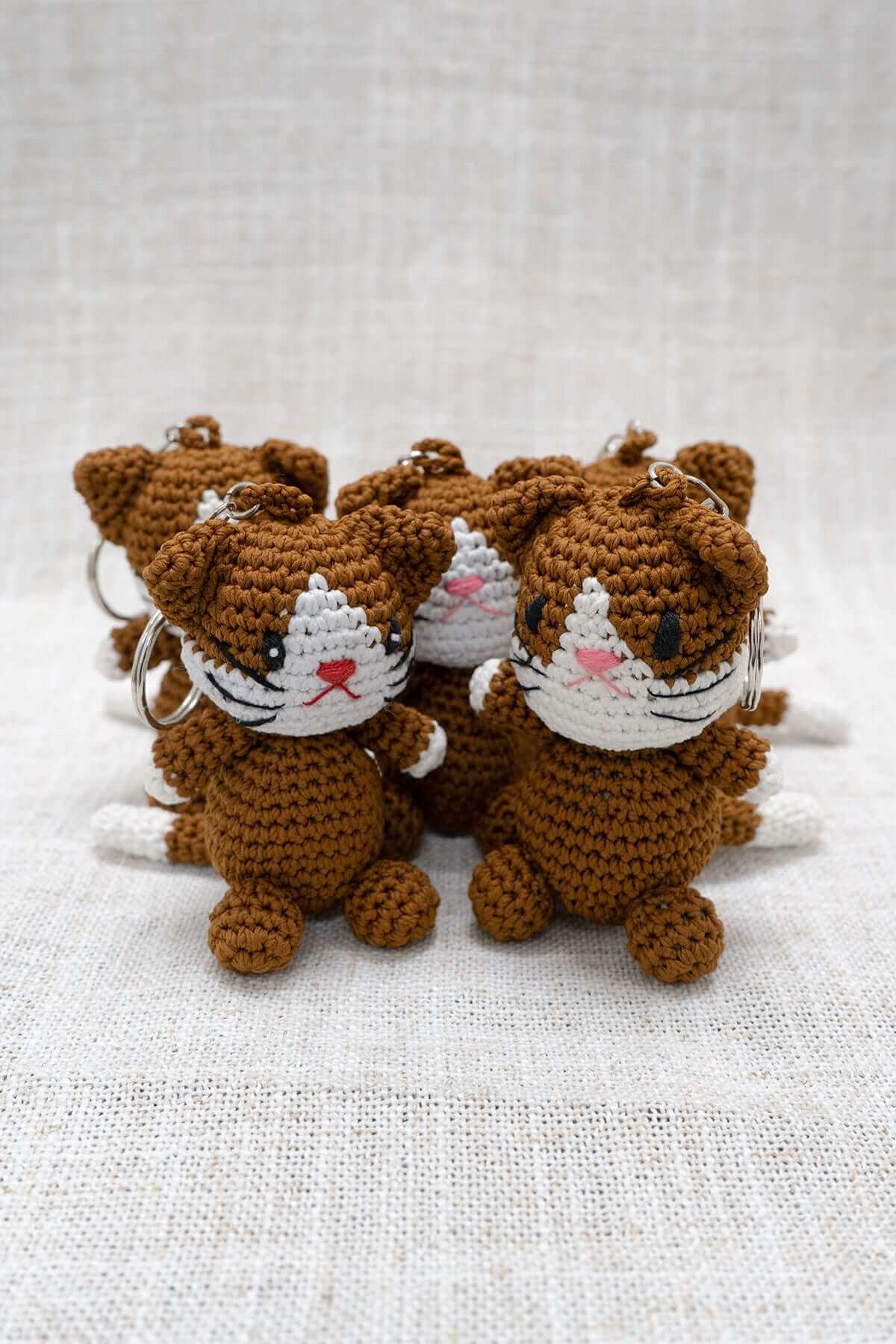Hand-crocheted brown and white cat keychains displayed together on a neutral fabric surface